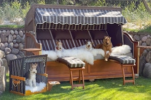 Un área de estar al aire libre con muebles de madera, un toldo a rayas, un muro de piedra y una caseta para perros con techo blanco y esponjoso.