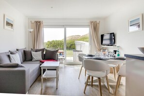 A modern living room with a grey sofa, a dining table set with glasses and plates, a TV mounted on the wall, and a view of greenery through the window. - An der Seedüne App. 1 - In Bester, Strandnaher Lage Wenningstedts Gelegen (Wenningstedt-Braderup (Sylt))
