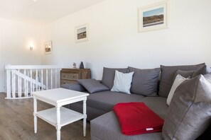 A modern living room with a grey sofa, a white coffee table, and a red throw blanket. There is a staircase with white railings and a wooden dresser in the background. - An der Seedüne App. 1 - In Bester, Strandnaher Lage Wenningstedts Gelegen (Wenningstedt-Braderup (Sylt))