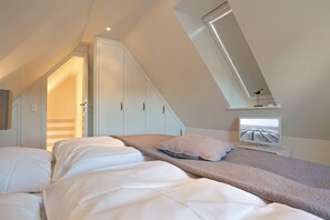 A bedroom with a sloped ceiling, a bed with white and beige bedding, a small desk with a computer, and a window with blinds. - Maisonette No. 8 - Erleben Sie Einen der Beliebtesten Urlaubsorte der Insel Sylt - Wenningstedt (Wenningstedt-Braderup (Sylt))