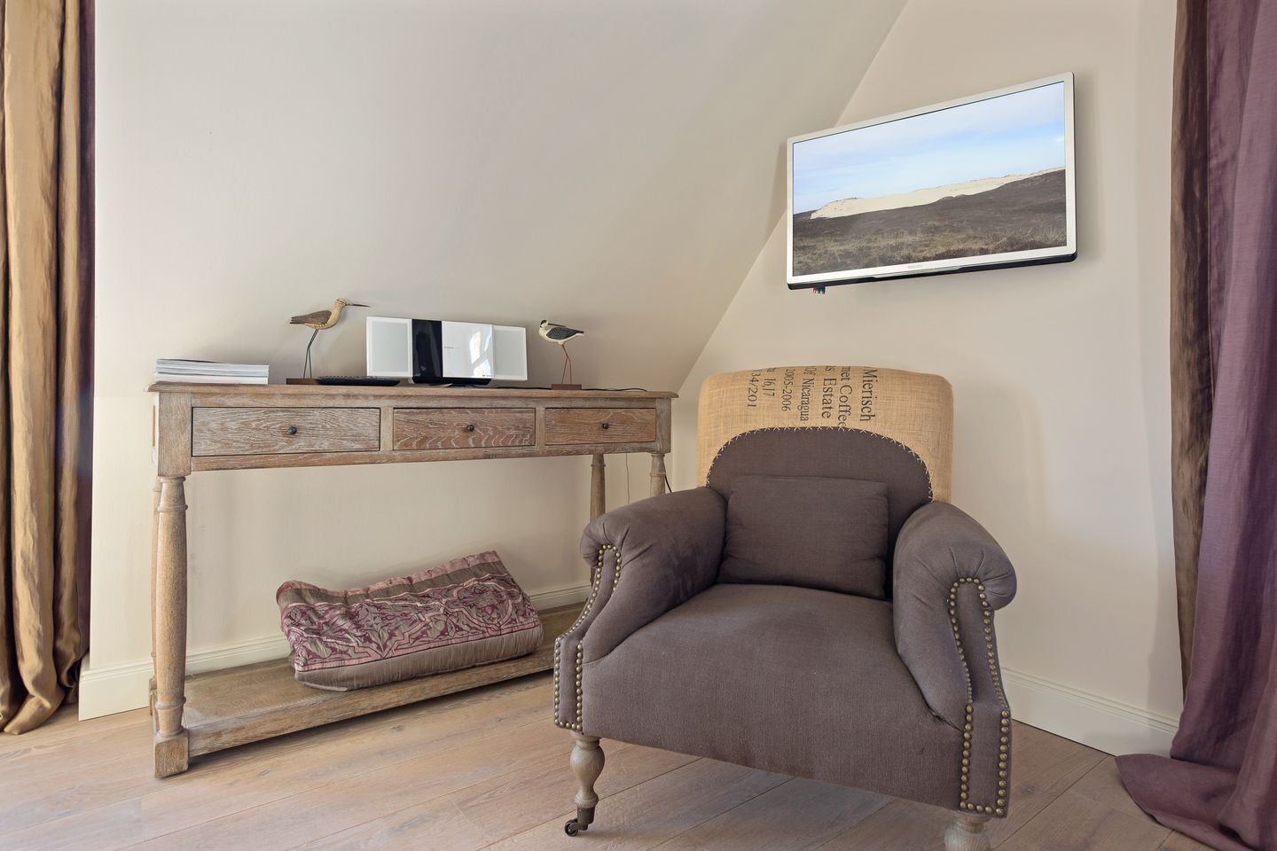 A cozy room with a wooden desk, a comfortable armchair, a cushioned bench, and a framed landscape painting on the wall.