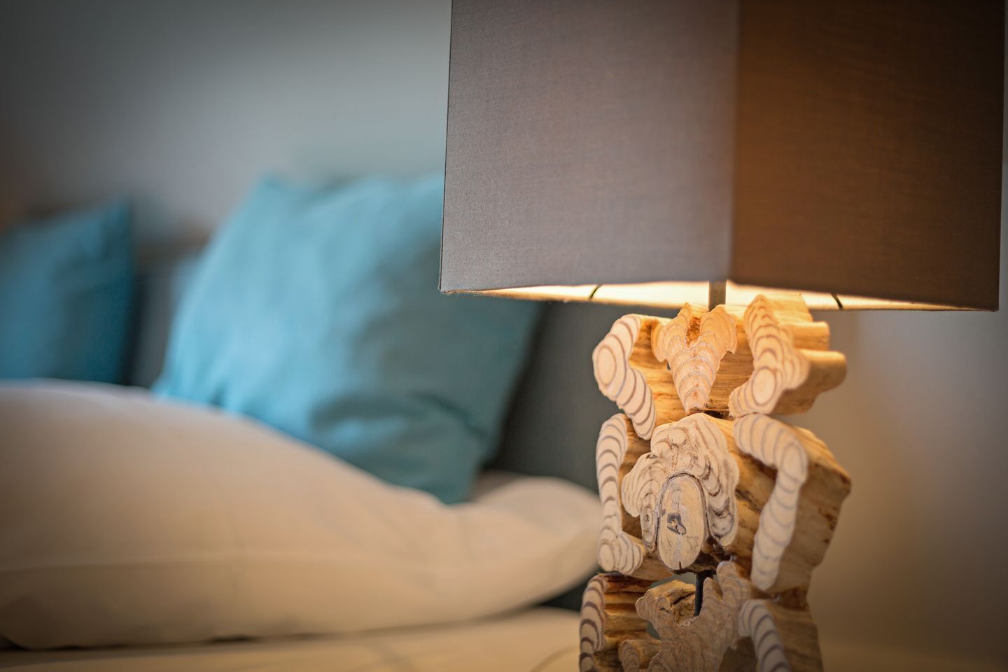Ein Bett mit weißer Bettwäsche und blauen Kissen, eine hölzerne Lampe mit außergewöhnlichem Design und ein dunkel gefärbter Lampenschirm.