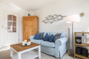 A living room with a light blue sofa, a wooden cabinet, a white table with candles, a floor lamp, and a JVC speaker system. - Kaptein Hein Cors, App. 5 - Llichtdurchfluteten Feriendomizil in Wenningstedt für bis zu 4 Personen (Wenningstedt-Braderup (Sylt))