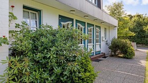 Ein zweistöckiges Gebäude mit weißer Fassade und türkisfarbenen Akzenten, mit einer kleinen Veranda und einem gepflegten Garten.