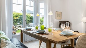 Un coin repas avec une table en bois dressée pour deux personnes, une vue sur le jardin grâce à de grandes fenêtres, et un espace salon confortable avec un canapé et des fauteuils.