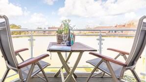 Ein Holztisch mit zwei Stühlen, einer Flasche und einem Glas auf einem Balkon mit Geländer.
