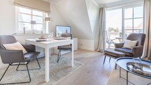 Ein modernes Wohnzimmer mit einem weißen Tisch, zwei grauen Sesseln, einem kleinen runden Tisch mit einem Glas und Blick auf Gebäude durch große Fenster.