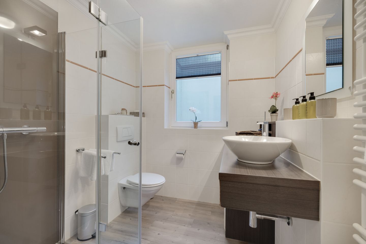 A modern bathroom with a glass shower enclosure, a white toilet, a large sink with a wooden countertop, and a window with a view of the sky.