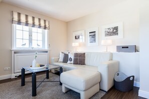 Un salon moderne avec un canapé blanc, une table basse en verre et un radiateur. Trois tableaux encadrés ornent le mur, et un petit haut-parleur noir est posé sur une étagère.