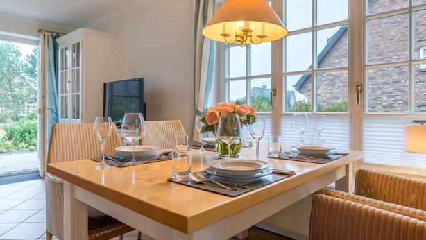 A dining area with a large table set for four, surrounded by wicker chairs, a chandelier, and a view of a brick building through large windows. - Heide & Meer, App. 4 - Sylt. Heide. Und Meer (Wenningstedt-Braderup (Sylt))