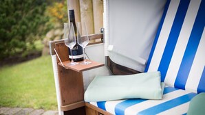 A blue and white striped lounge chair with a cushion, a small wooden table with two wine glasses and a bottle, and a garden view. - Heide & Meer, App. 4 - Sylt. Heide. Und Meer (Wenningstedt-Braderup (Sylt))