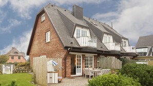 A two-story house with a brick facade, a sloped roof, and a wooden deck with outdoor furniture. - Heide & Meer, App. 4 - Sylt. Heide. Und Meer (Wenningstedt-Braderup (Sylt))