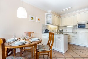 A modern kitchen with white cabinetry, a central island, and a dining area with a wooden table and chairs. - Haus Carina, App. 55 - 3-raum Appartement im Erdgeschoss und Untergeschoss mit ca. 65m² (Wenningstedt-Braderup (Sylt))