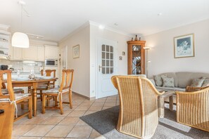 A well-lit living room with a sofa, two wicker chairs, a dining table with chairs, a kitchen area, and a door with a glass panel. - Haus Carina, App. 55 - 3-raum Appartement im Erdgeschoss und Untergeschoss mit ca. 65m² (Wenningstedt-Braderup (Sylt))