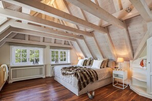A bedroom with a sloped ceiling, wooden beams, a large bed, two windows, a radiator, and a bedside table with a lamp. - Familier Hygge Hus - Außergewöhnliche Reetdachvilla am Ortsrand von Braderup (Wenningstedt-Braderup (Sylt))