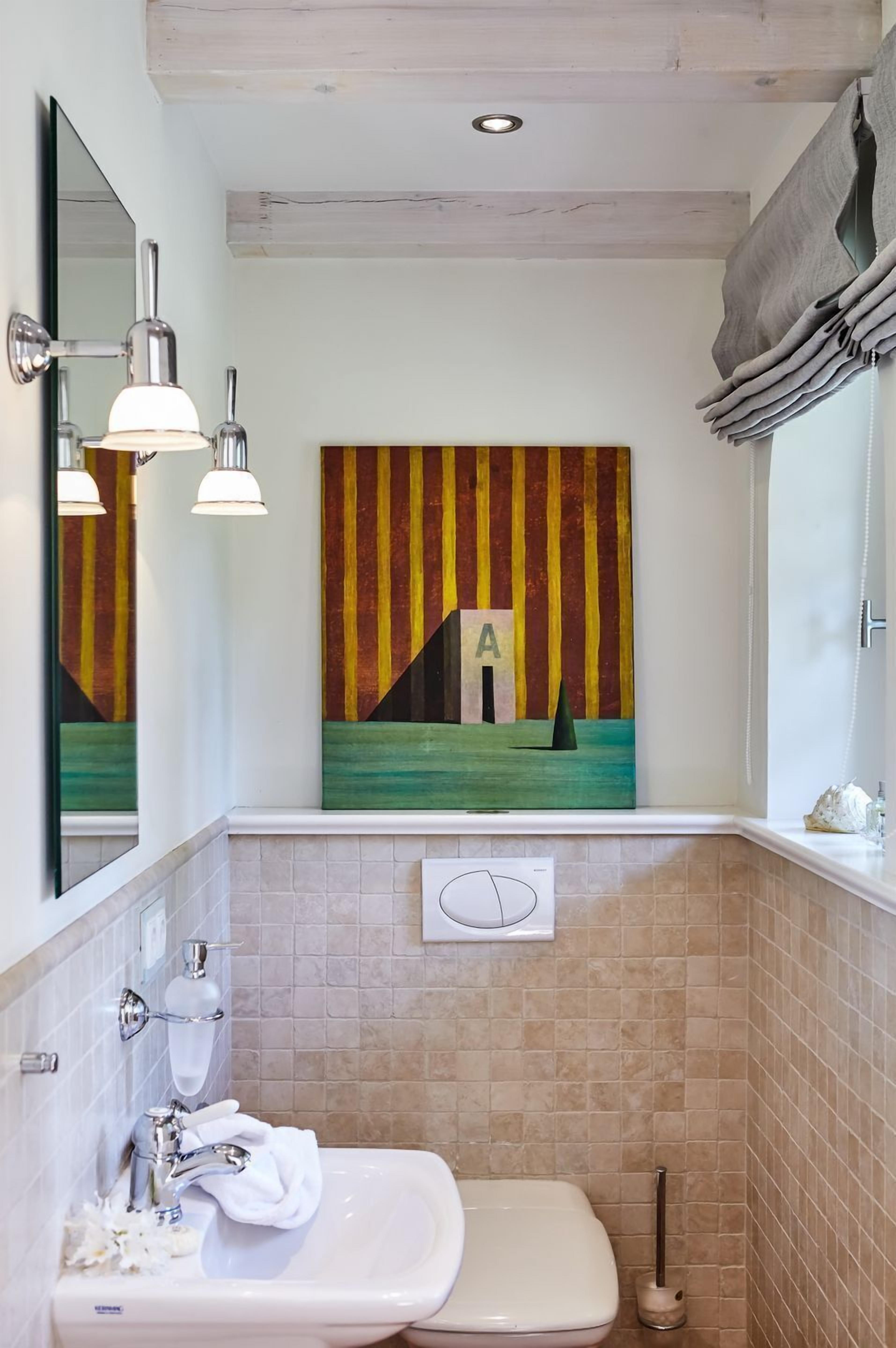Une salle de bain avec un lavabo blanc, un miroir et un tableau représentant une maison accroché au mur.