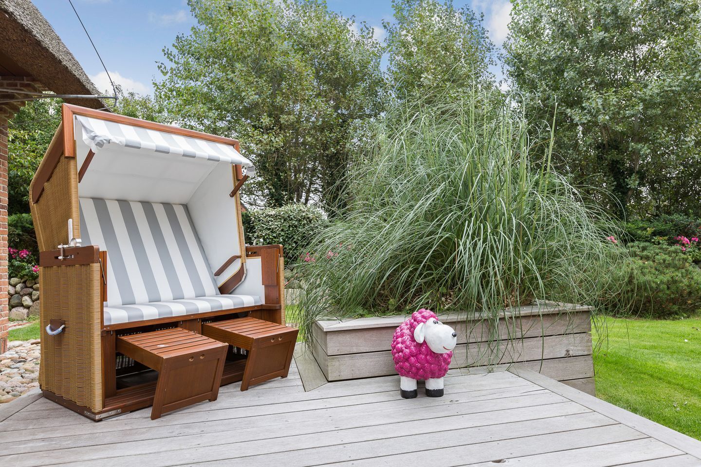 A wooden deck with a wicker lounge chair and a small table, surrounded by greenery and a decorative sheep statue.