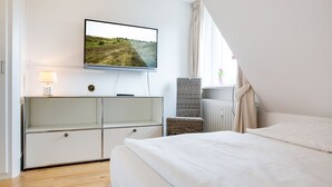 A bedroom with a bed, a TV mounted on the wall, a dresser, a chair, and a window with curtains. - Syltblume, App. 11 - Viel Urlaub in Lauschiger Lage (Wenningstedt-Braderup (Sylt))