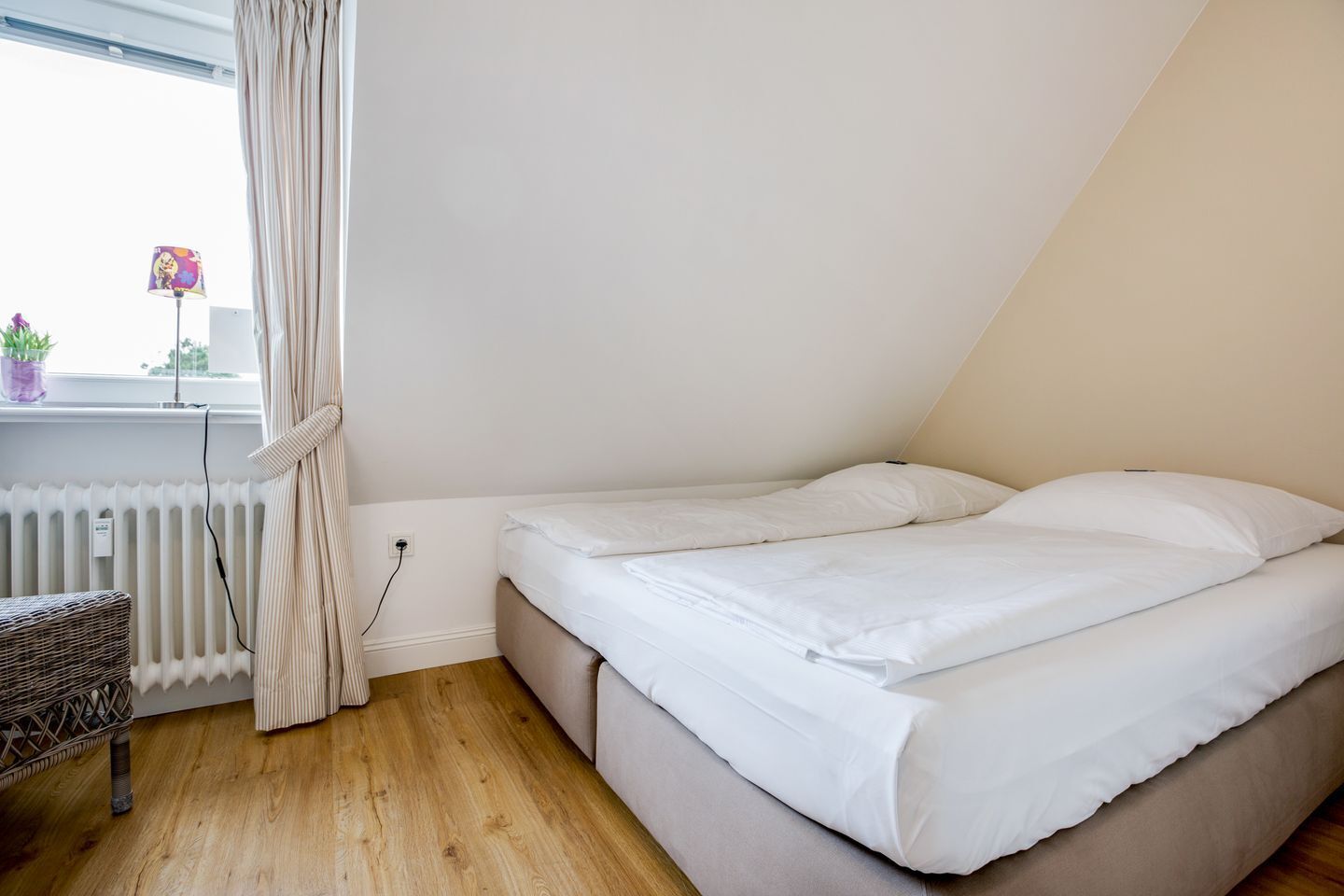 A small, single-bed room with a sloped ceiling, a radiator, a wicker chair, and a window with curtains.