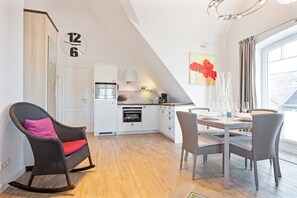 Une cuisine moderne avec des meubles blancs, un coin repas avec une table dressée pour deux personnes, et une grande fenêtre avec des rideaux.