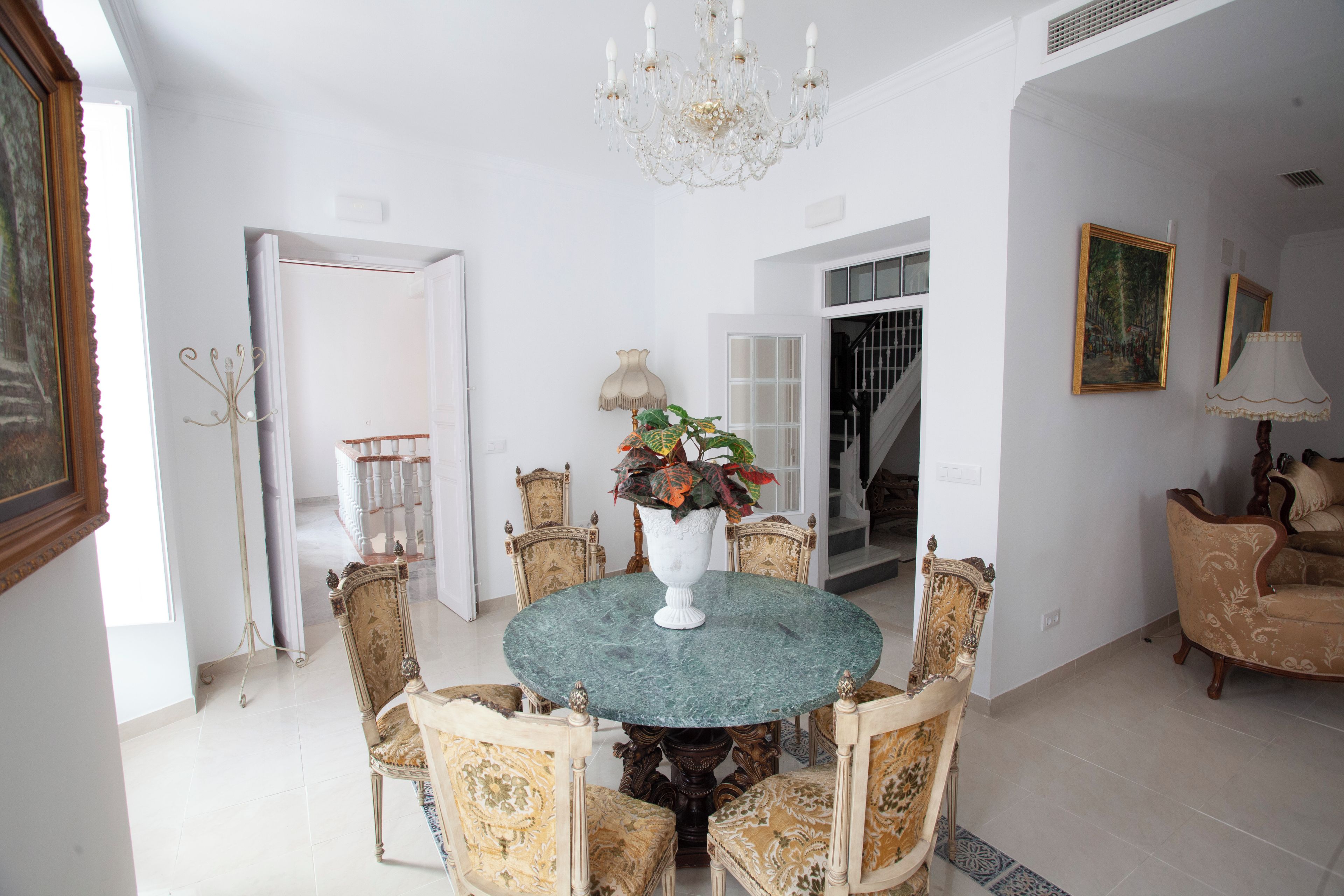 Un comedor con una mesa redonda rodeada de sillas ornamentadas, una araña y un jarrón con flores.