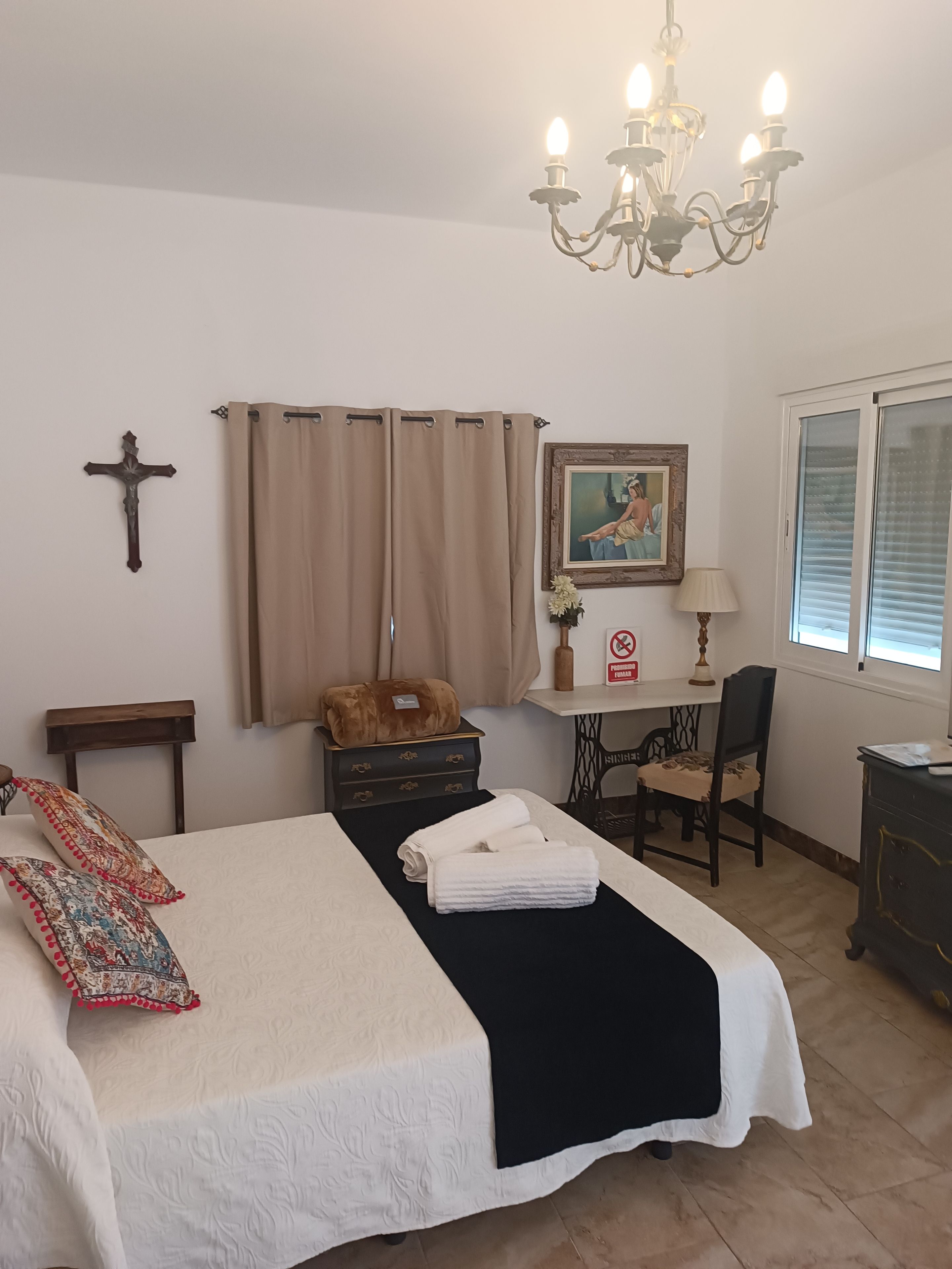 Un dormitorio ordenado con cama, escritorio, silla, una cruz en la pared y una ventana con persianas.