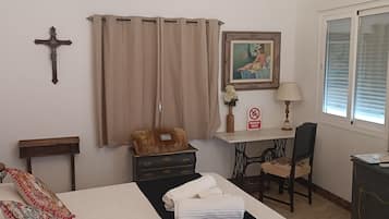 Un dormitorio ordenado con cama, escritorio, silla, una cruz en la pared y una ventana con persianas.