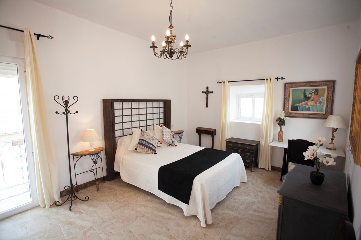 Quarto com cama, mesas de cabeceira, escrivaninha, cadeira, cruz na parede e lustre.