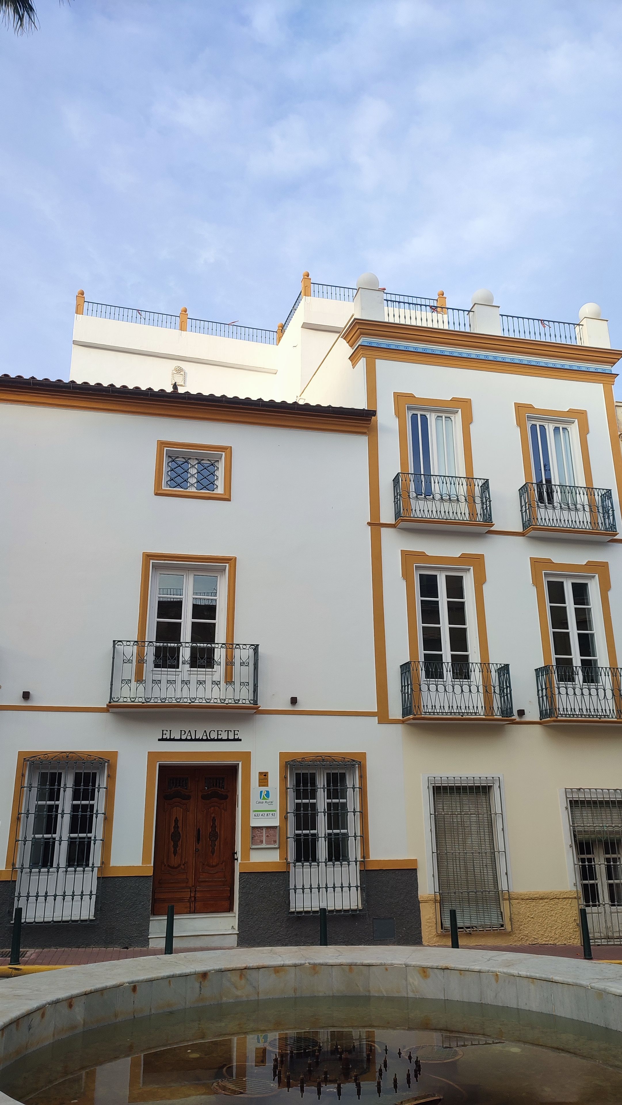 Un edificio con varios balcones, una entrada central y un patio con una piscina reflectante.