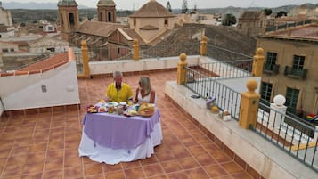 A couple dining on a terrace with a table set for two, overlooking a cityscape with historic buildings and a clear sky.