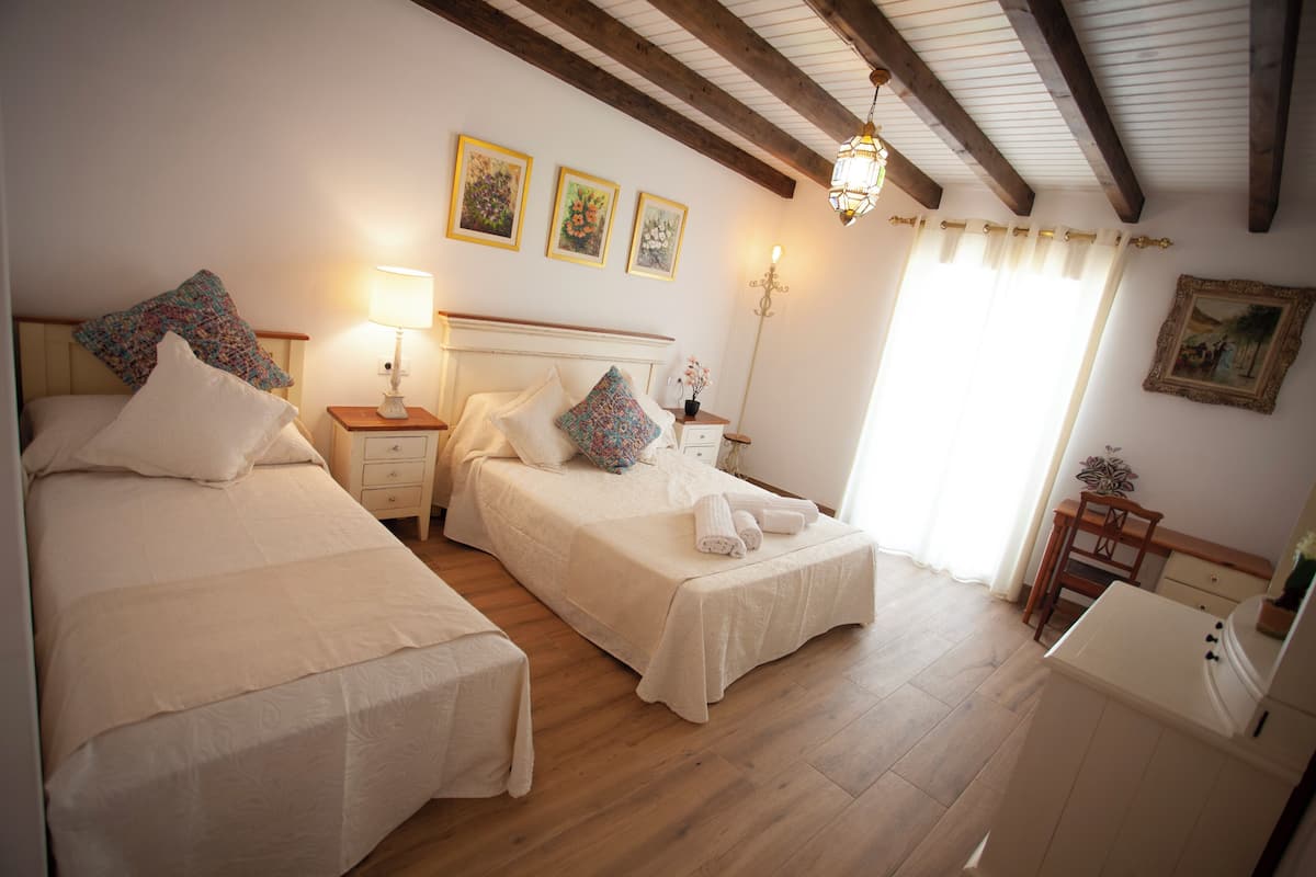 Ein Schlafzimmer mit zwei Betten, einem Nachttisch mit Lampe, einem Schreibtisch und einem Stuhl. An der Wand hängen drei gerahmte Bilder und eine Chandelüster.