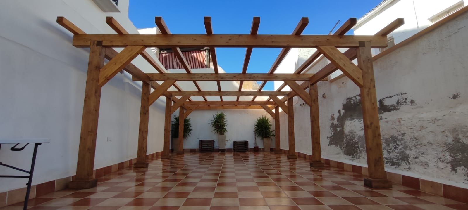 Zona exterior coberta amb una pèrgola de fusta, sòl de rajola i plantes en testos.