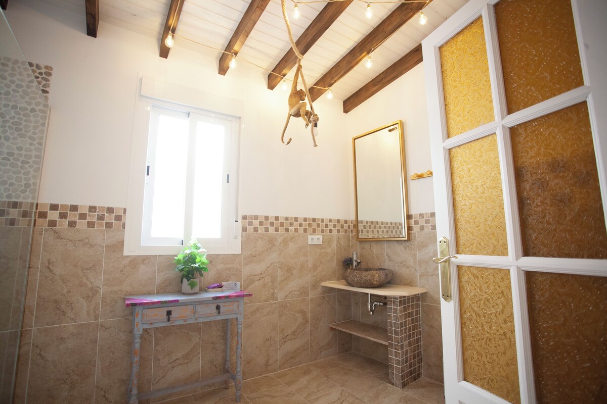 Un baño con pared de baldosas de piedra, techo de madera, ventana, lavamanos con lavabo de piedra y espejo con marco dorado.