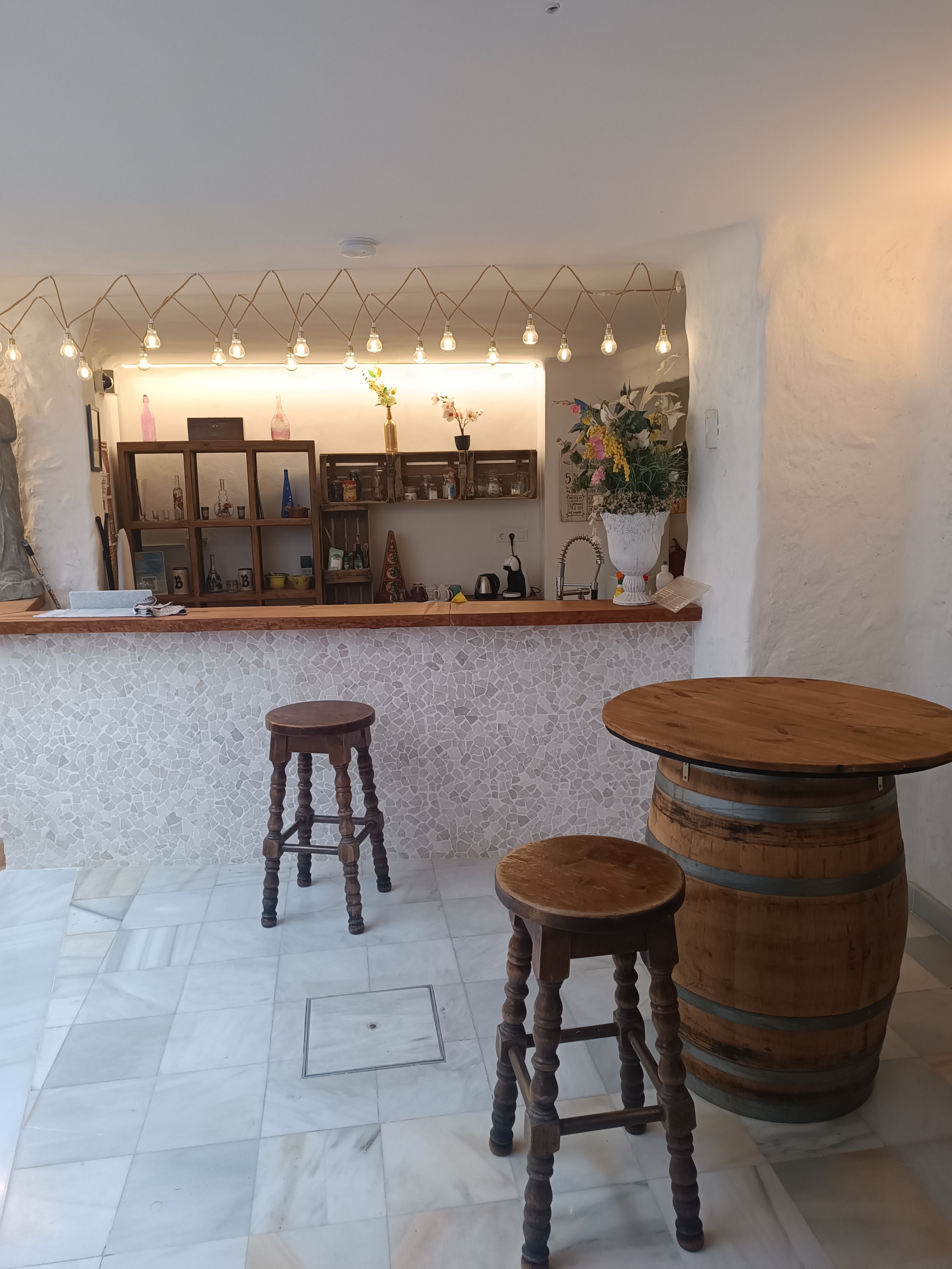 Zona de bar con suelo de mosaico, taburetes de madera, una mesa redonda de madera y una barra con estantes y objetos decorativos.