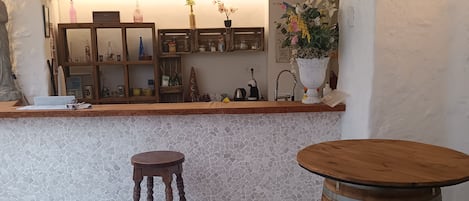 Ένας χώρος μπαρ με δάπεδο επενδεδυμένο με μωσαϊκά, ξύλινα σκαμπό, ένα στρογγυλό ξύλινο τραπέζι και έναν πάγκο μπαρ με ράφια και διακοσμητικά αντικείμενα.