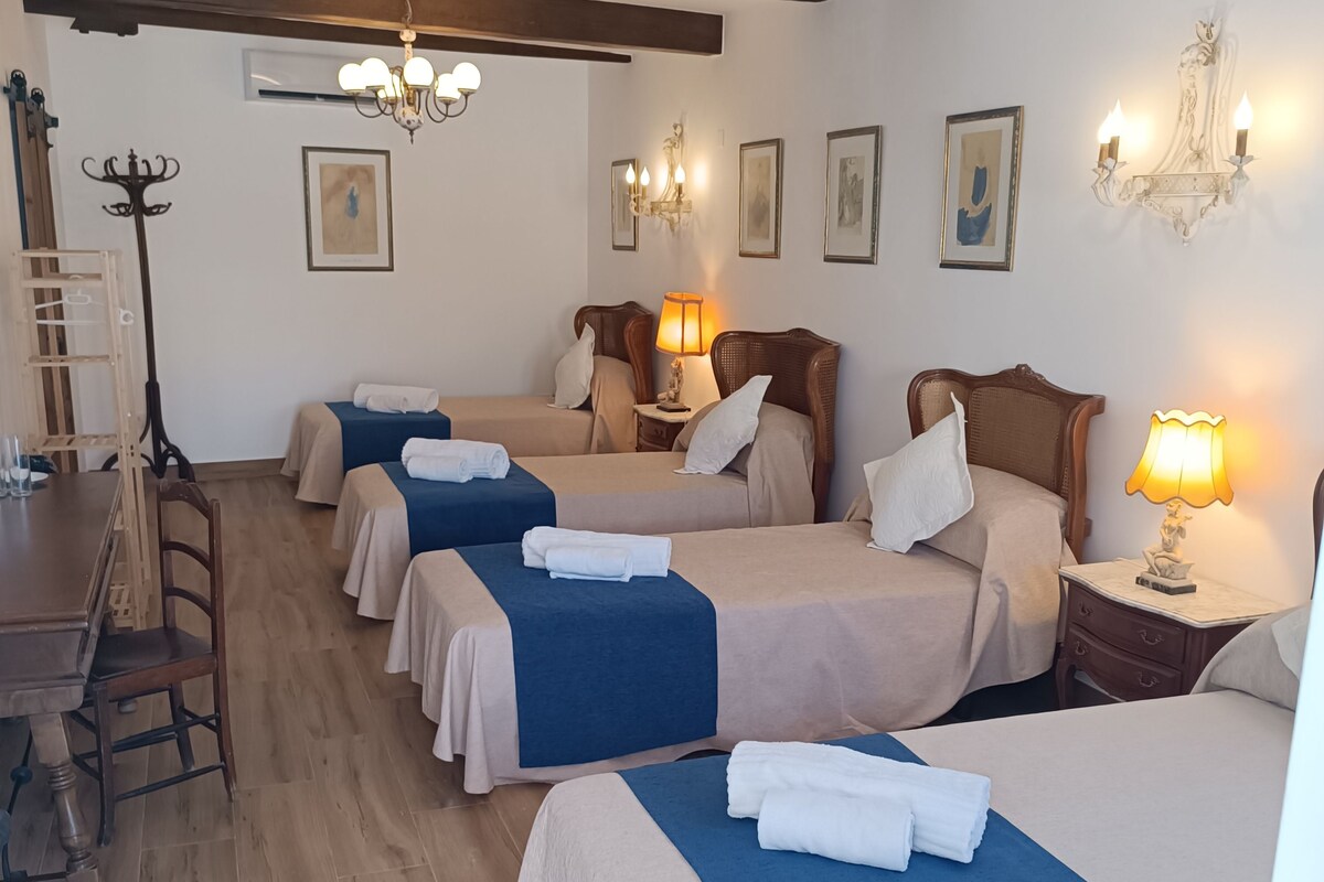 Quarto de hotel com quatro camas, piso de madeira, uma mesa de jantar e luminárias fixadas na parede.