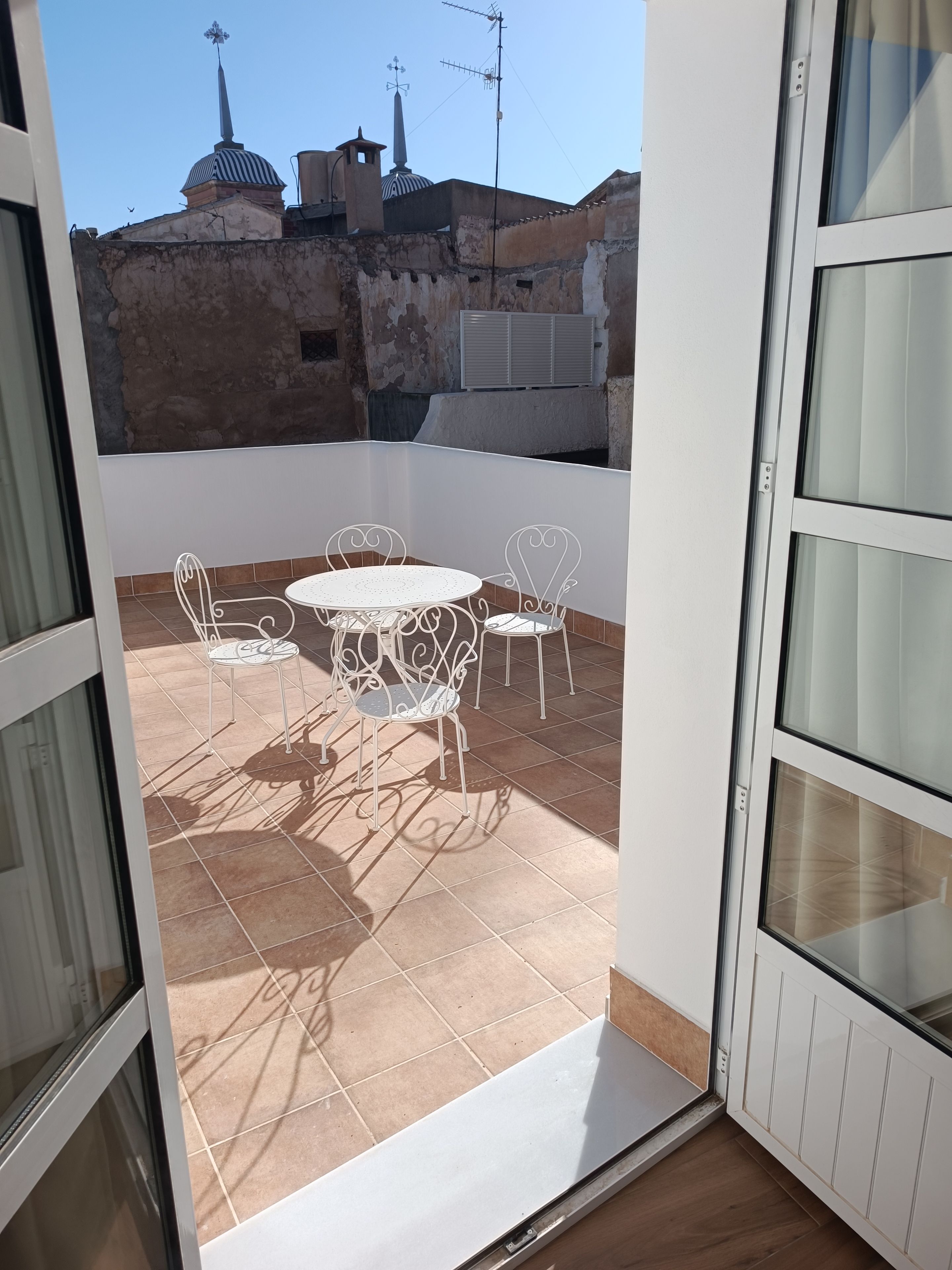 Un balcón abierto con una mesa y sillas de metal blanco, suelo de baldosas y vistas a edificios con cúpulas y antenas al fondo.