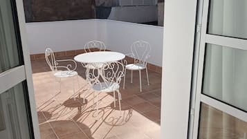 Un balcón abierto con una mesa y sillas de metal blanco, suelo de baldosas y vistas a edificios con cúpulas y antenas al fondo.