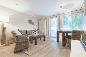 A spacious living room with a sofa, a coffee table, a dining table with chairs, a large window with curtains, and a decorative wall piece. - Mer-du-nord - Mer-du-nord - Wunderschöne Wohnung im Riviera-landhausstil (Sylt)