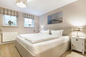 A bedroom with a large bed, two bedside tables, a radiator, and a window with curtains. - Mer-du-nord - Mer-du-nord - Wunderschöne Wohnung im Riviera-landhausstil (Sylt)