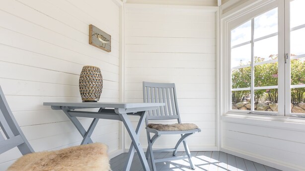 A corner room with a white wooden wall, a table with a woven vase, two chairs with fur cushions, and a window with a view of greenery. - Sylter Quartier, Whg. 1 - In Zentraler und Dennoch Ruhiger Lage Westerlands Gelegenes 2-raum (Sylt)