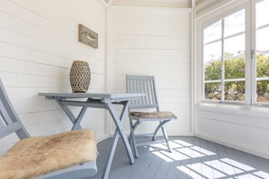 A corner room with a white wooden wall, a table with a woven vase, two chairs with fur cushions, and a window with a view of greenery. - Sylter Quartier, Whg. 1 - In Zentraler und Dennoch Ruhiger Lage Westerlands Gelegenes 2-raum (Sylt)
