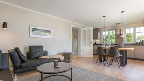A modern living room with a sofa, a coffee table, and a dining area with chairs and a kitchen in the background. - Sylter Quartier, Whg. 1 - In Zentraler und Dennoch Ruhiger Lage Westerlands Gelegenes 2-raum (Sylt)
