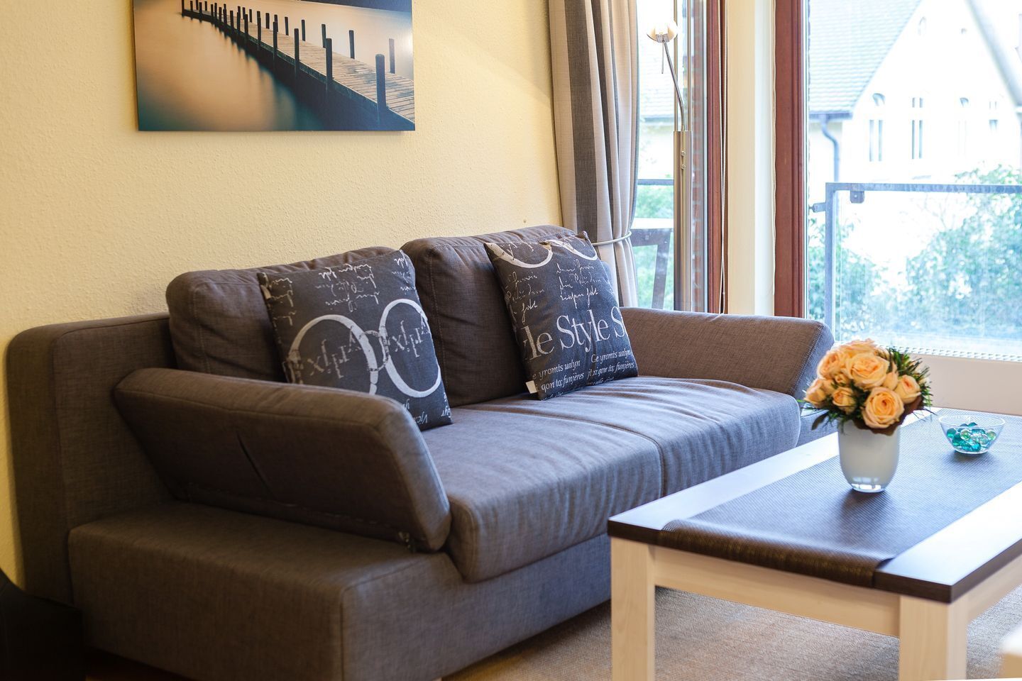 A modern living room with a grey sofa, two decorative pillows, a small wooden coffee table, and a vase with flowers.