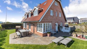 A two-story house with a red-tiled roof, a patio with outdoor furniture, and a fenced backyard. - Haus am Strand, Beachfront - Strand, Meer & Luxus (Wenningstedt-Braderup (Sylt))