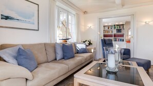 A living room with a beige sofa, a glass coffee table, and a bookshelf filled with books. - Haus am Strand, Beachfront - Strand, Meer & Luxus (Wenningstedt-Braderup (Sylt))