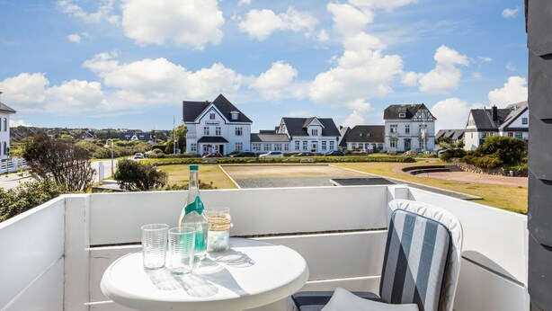 A balcony with a table, a bottle, and glasses, overlooking a row of white houses with dark roofs. - Am Kurpark, App. 4 - Wohlfühl-appartement in Bester Lage. Nur Wenige Meter zum Strand (Wenningstedt-Braderup (Sylt))