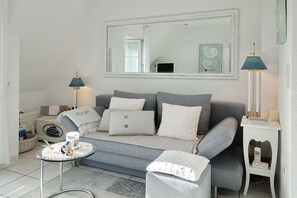 Una sala de estar moderna con un sofá gris, una mesa de centro redonda de vidrio, una mesita auxiliar blanca con una lámpara y un espejo grande en la pared.