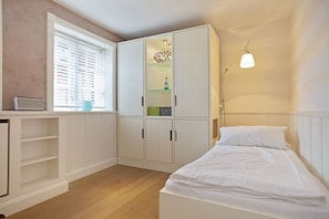 Un dormitorio con cama, armario, estante con objetos, ventana con persianas y lámpara.