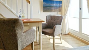 Ein Wohnzimmer mit einem Holztisch, zwei Sesseln, einem an der Wand befestigten Fernseher und einem großen Fenster mit Vorhängen.